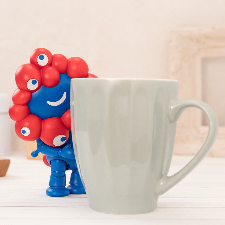 Colorful figurine with multiple eyes next to a white mug on a light wooden surface.