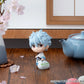 Small figurine with light blue hair next to a teapot and vase on a wooden surface.